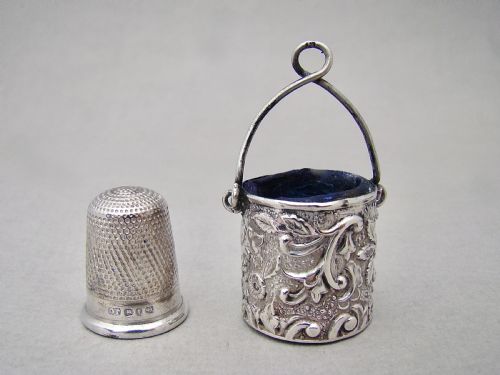 fig 1. A Victorian sterling silver bucket-shaped thimble holder with an arched wire handle and embossed floral repousse patterns, containing a silver thimble.