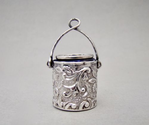 fig 2. A Victorian sterling silver bucket-shaped thimble holder with an arched wire handle and embossed floral repousse patterns, containing a silver thimble.