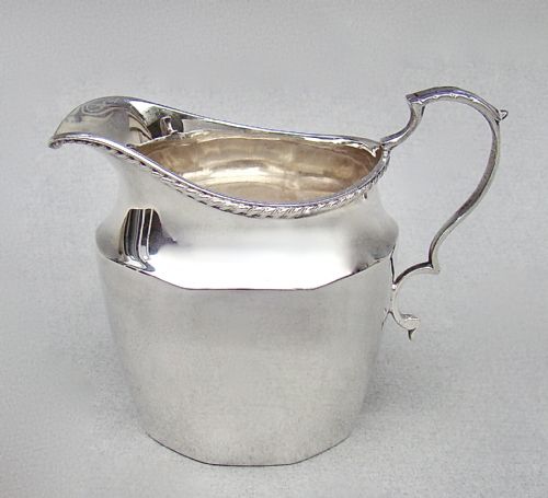 fig 1. A polished George V sterling silver cream jug featuring a faceted body, C-scroll handle, and stamped English hallmarks near the gadrooned rim.