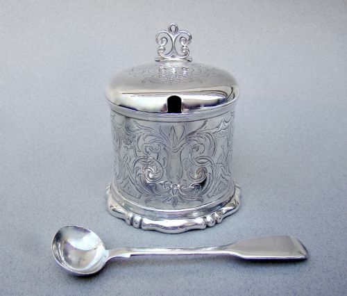 fig 2. A cylindrical Victorian sterling silver mustard pot decorated with intricate foliate scroll engraving, featuring a hinged lid, thumbpiece, scroll handle, and spoon.