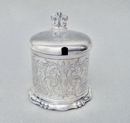 fig 1. A cylindrical Victorian sterling silver mustard pot decorated with intricate foliate scroll engraving, featuring a hinged lid, thumbpiece, scroll handle, and spoon.