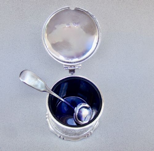 fig 3. A cylindrical Victorian sterling silver mustard pot decorated with intricate foliate scroll engraving, featuring a hinged lid, thumbpiece, scroll handle, and spoon.