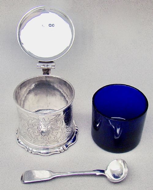 fig 4. A cylindrical Victorian sterling silver mustard pot decorated with intricate foliate scroll engraving, featuring a hinged lid, thumbpiece, scroll handle, and spoon.