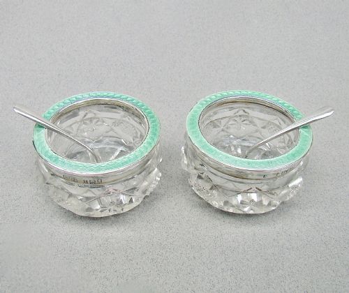 fig 1. A pair of circular cut glass salt cellars featuring sterling silver rims decorated with mint green guilloche enamel, shown alongside two small silver salt spoons.