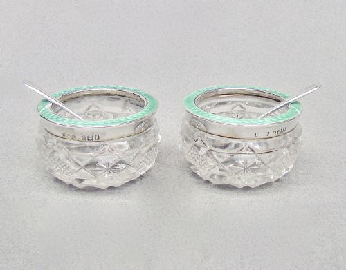 fig 2. A pair of circular cut glass salt cellars featuring sterling silver rims decorated with mint green guilloche enamel, shown alongside two small silver salt spoons.