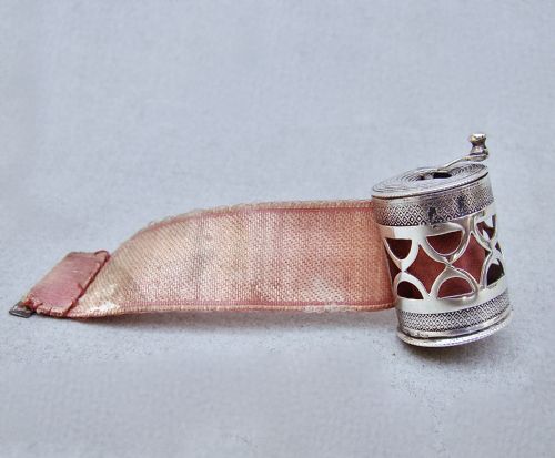 fig 1. A drum-shaped 19th-century Dutch silver tape measure featuring a pierced geometric casing, top winding handle, and extending pink silk ribbon.