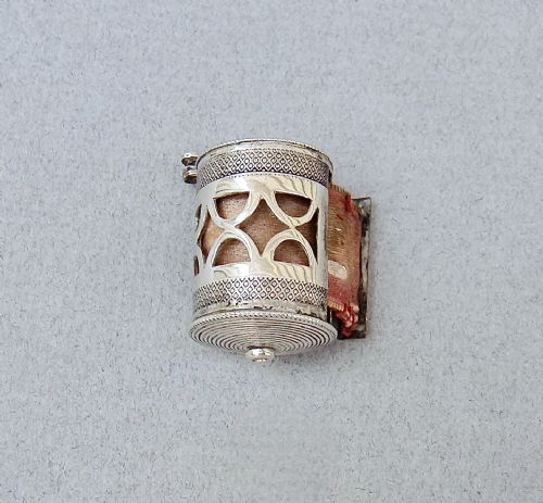 fig 2. A drum-shaped 19th-century Dutch silver tape measure featuring a pierced geometric casing, top winding handle, and extending pink silk ribbon.