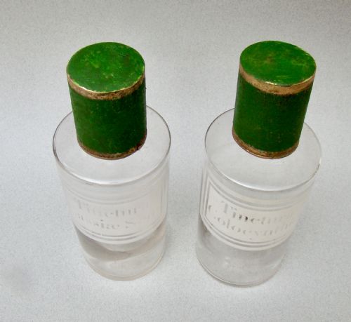 fig 1. A pair of clear glass 19th-century apothecary jars featuring white etched Latin labels and cylindrical green painted stoppers with metallic rims.