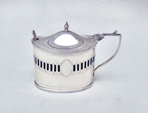 fig 1. An oval Edwardian sterling silver mustard pot featuring pierced geometric fretwork, an engraved cartouche, a hinged dome lid with a finial, and a side handle.