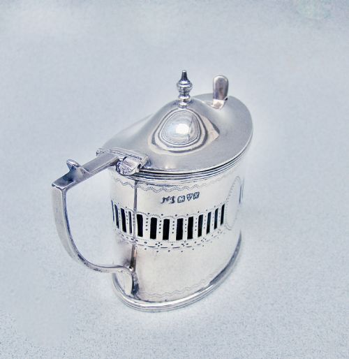 fig 3. An oval Edwardian sterling silver mustard pot featuring pierced geometric fretwork, an engraved cartouche, a hinged dome lid with a finial, and a side handle.