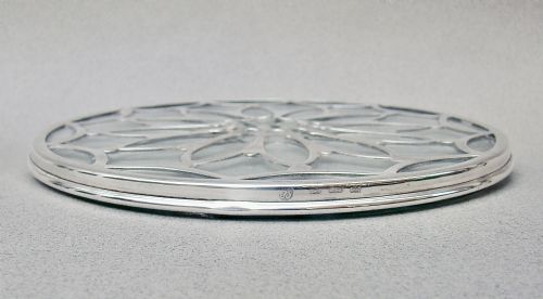 fig 1. A circular clear glass teapot stand featuring an intricate pierced sterling silver overlay in a petalled geometric pattern.