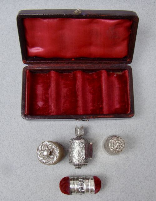 fig 3. An open dark-cased Victorian sewing set lined with crimson red silk velvet, containing four decorated silver tools including a thimble, needle case, pin cushion, and waxer.