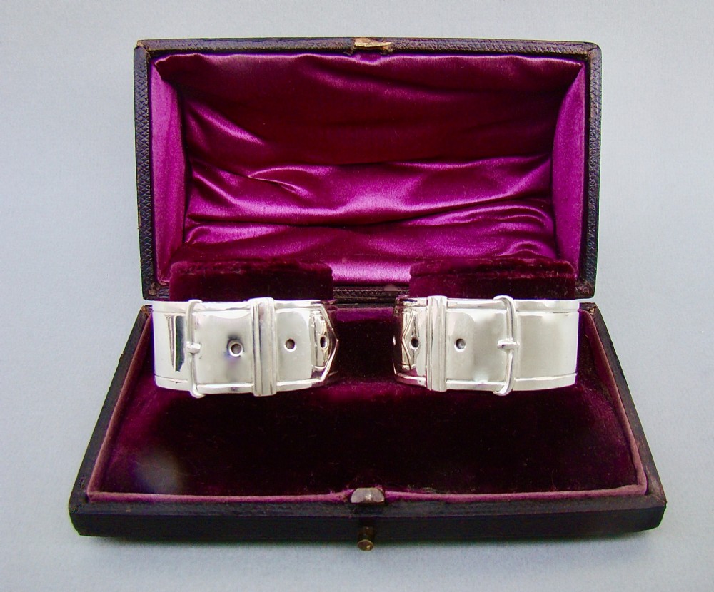 A pair of Victorian silver-plated napkin rings designed to resemble buckled belts, resting inside a velvet-lined presentation box. View II