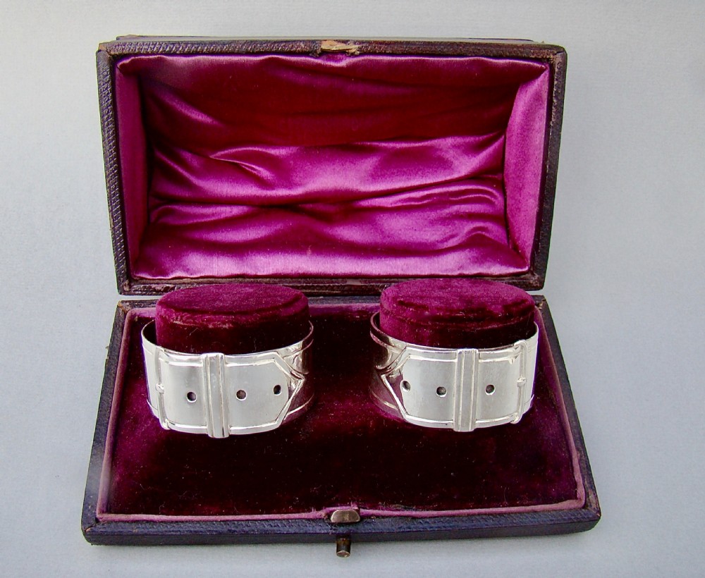 A pair of Victorian silver-plated napkin rings designed to resemble buckled belts, resting inside a velvet-lined presentation box.