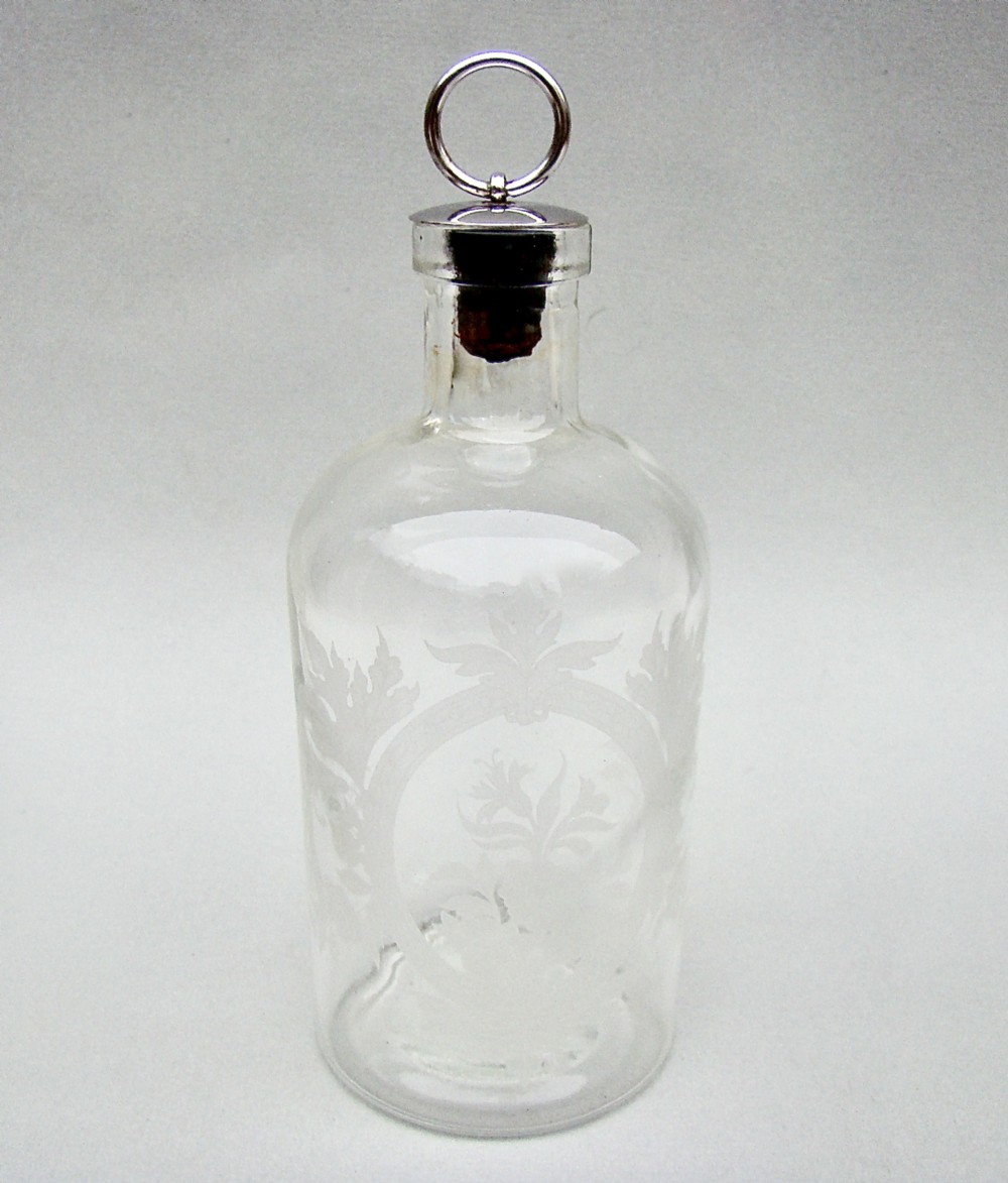 A clear glass cologne bottle with acid-etched floral patterns and a metal-capped cork stopper topped with a silver-coloured ring pull.