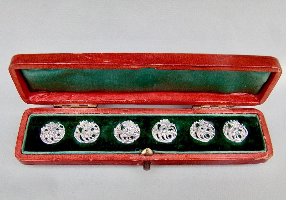A cased set of six Art Nouveau pierced sterling silver buttons arranged inside a red velvet-lined fitted box.