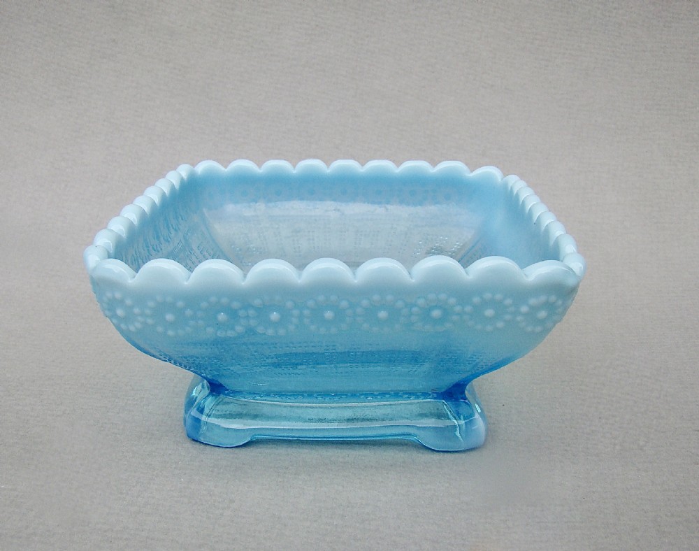 A square blue opalescent pressed glass bowl featuring a scalloped rim and moulded daisy pattern on a footed base.