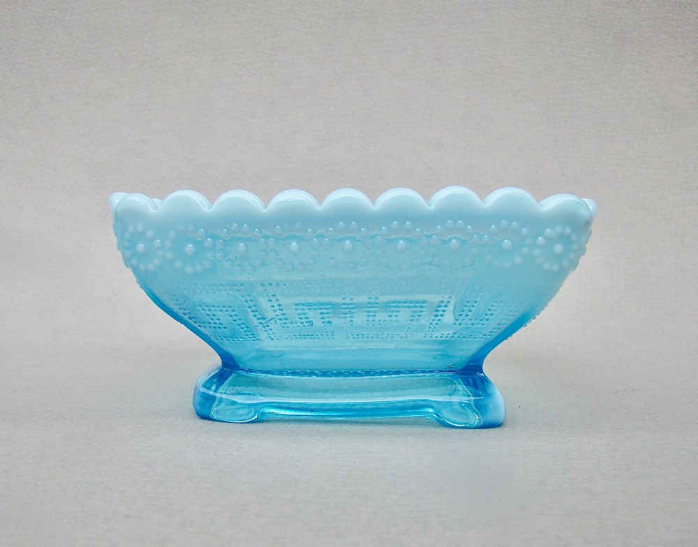 A square blue opalescent pressed glass bowl featuring a scalloped rim and moulded daisy pattern on a footed base. View II