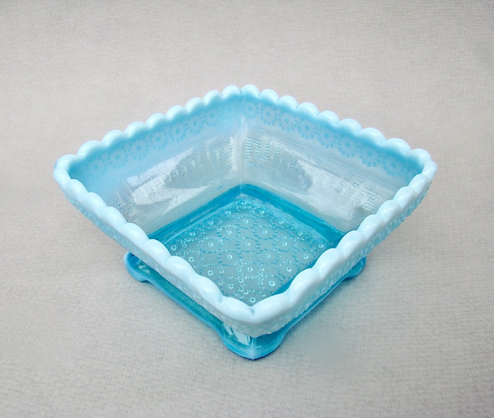 A square blue opalescent pressed glass bowl featuring a scalloped rim and moulded daisy pattern on a footed base. View III