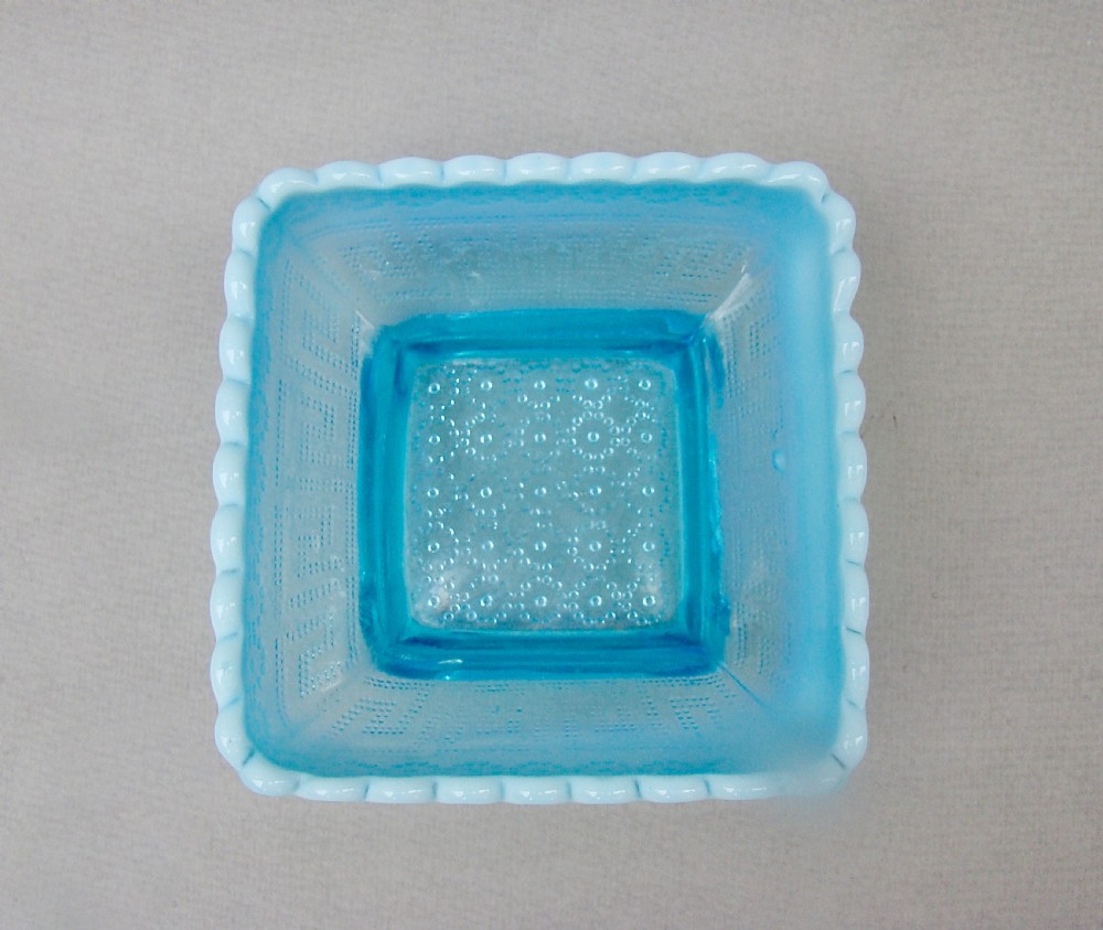 A square blue opalescent pressed glass bowl featuring a scalloped rim and moulded daisy pattern on a footed base. View IV