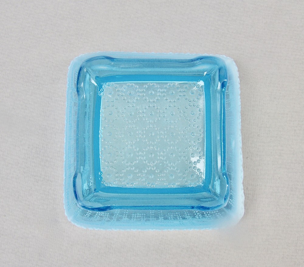 A square blue opalescent pressed glass bowl featuring a scalloped rim and moulded daisy pattern on a footed base. View V