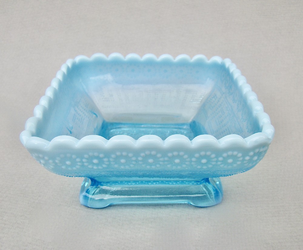A square blue opalescent pressed glass bowl featuring a scalloped rim and moulded daisy pattern on a footed base. View VII
