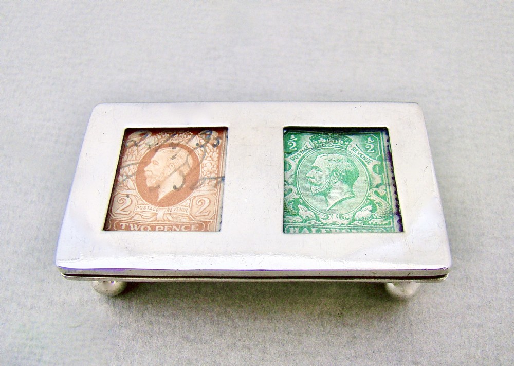 An Edwardian sterling silver double trough stamp box featuring twin glass apertures displaying British stamps, resting on four spherical feet.