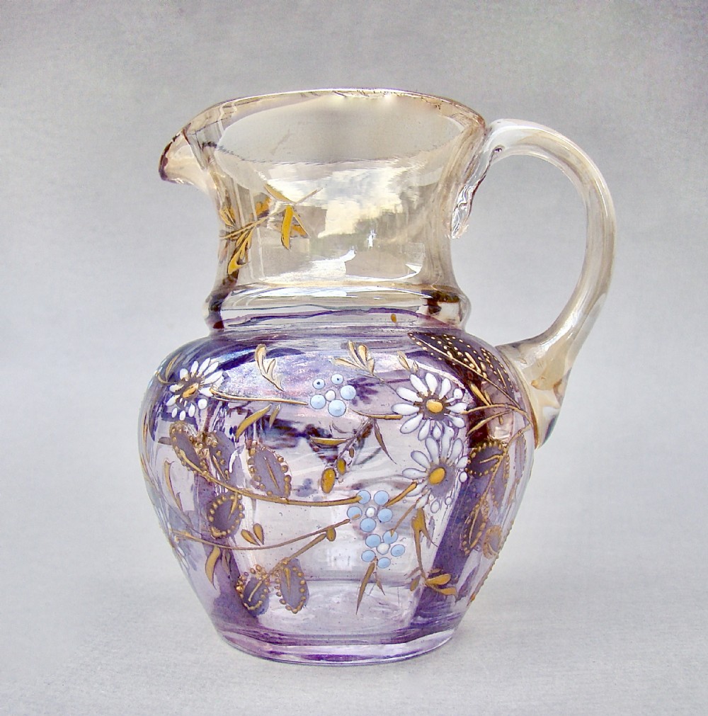 A late Victorian glass cream jug featuring purple and clear iridescent glass decorated with hand-painted white and gold enamelled floral motifs and an applied clear handle.