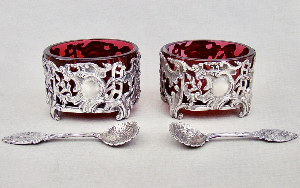 A pair of 19th-century French silver salt cellars with pierced rococo scrollwork frames, ruby glass liners, and matching silver salt spoons.