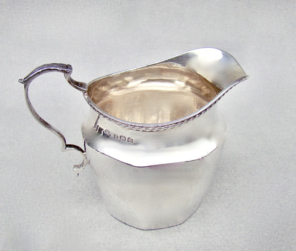 A polished George V sterling silver cream jug featuring a faceted body, C-scroll handle, and stamped English hallmarks near the gadrooned rim.