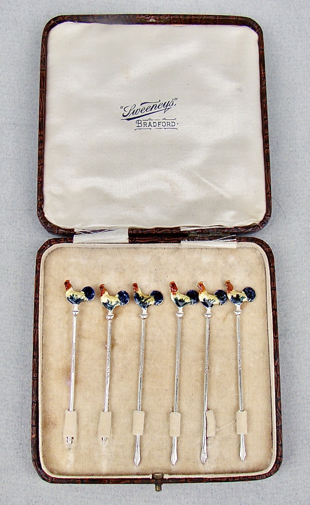 A cased set of six sterling silver cocktail sticks featuring colourful enamelled cockerel finials arranged inside a fitted cream-lined presentation box.
