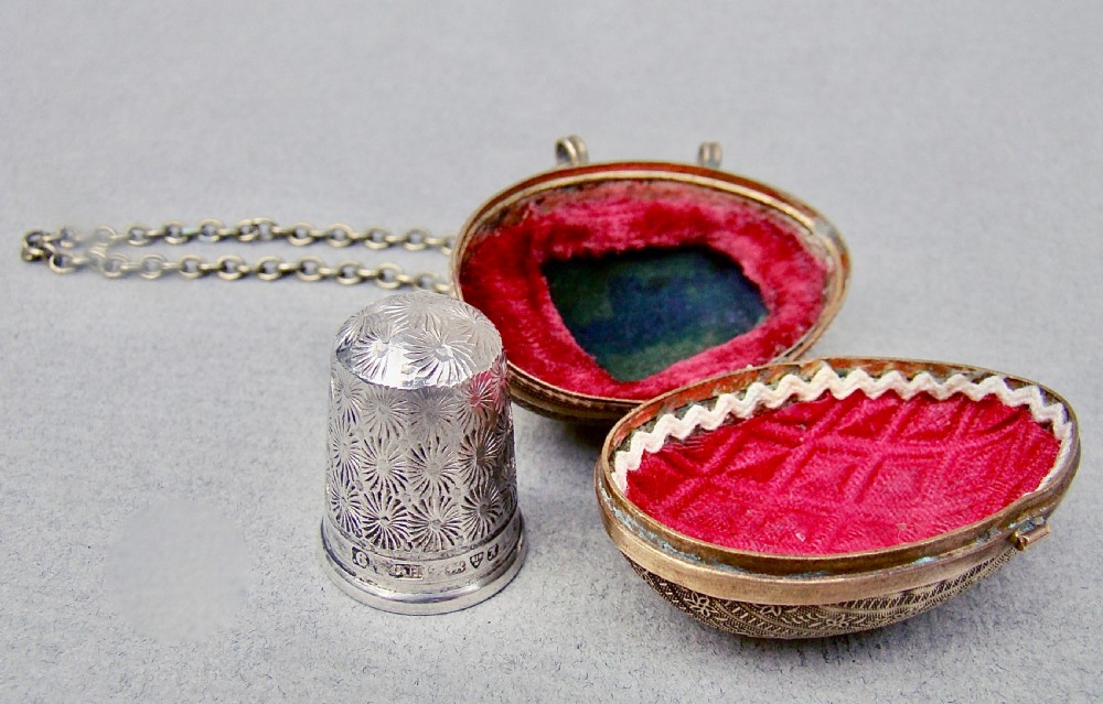 A hallmarked silver thimble with starburst engraving positioned beside an open egg-shaped ormolu case lined with red velvet.