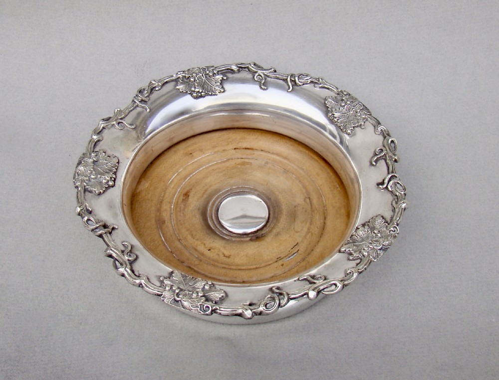 A circular William IV Old Sheffield Plate wine coaster featuring a turned wooden insert, central silver boss, and a chased border decorated with grapevines and leaves.