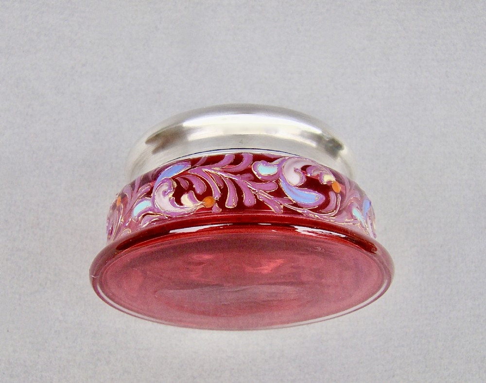 A small circular cranberry glass trinket box decorated with purple and blue enamelling, topped with a plain polished sterling silver lid. View IV