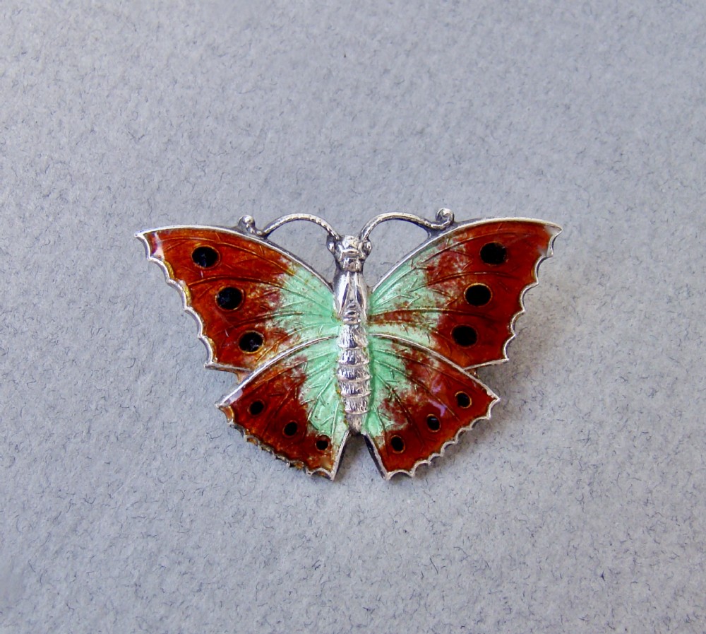 A 1930s sterling silver butterfly brooch featuring engine-turned guilloche enamel in shades of burnt orange and pale green with black spot details.