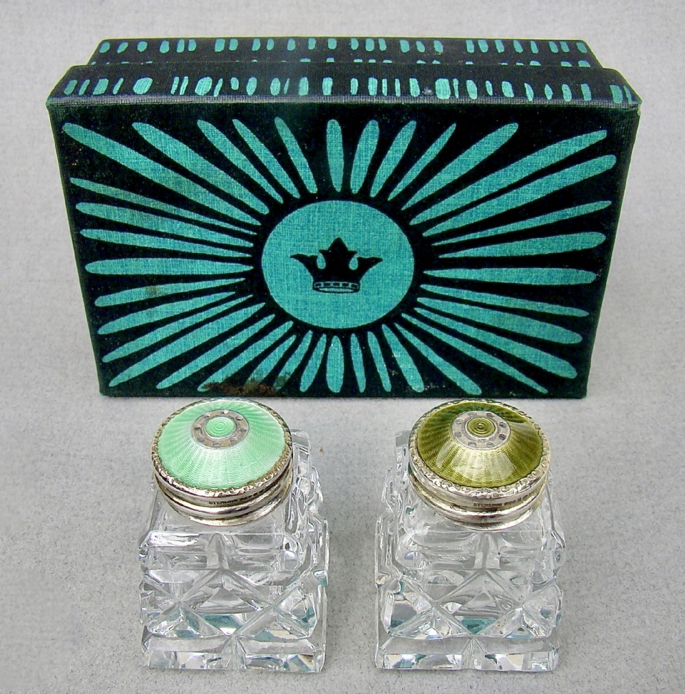 A pair of Art Deco cut glass salt shakers featuring green guilloche enamel and silver tops, shown in front of their fitted presentation box.
