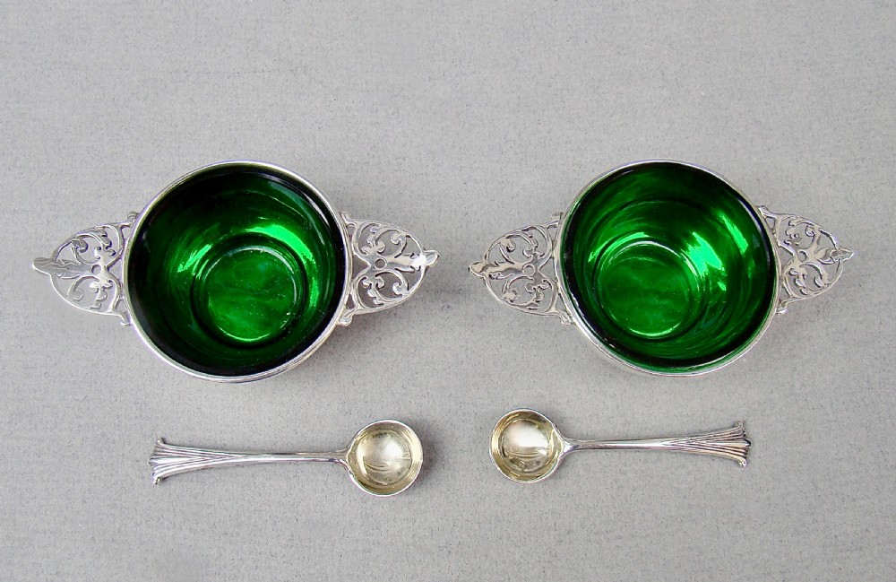A pair of sterling silver quaich-form open salt cellars with pierced handles, vivid green glass liners, and two matching silver salt spoons.