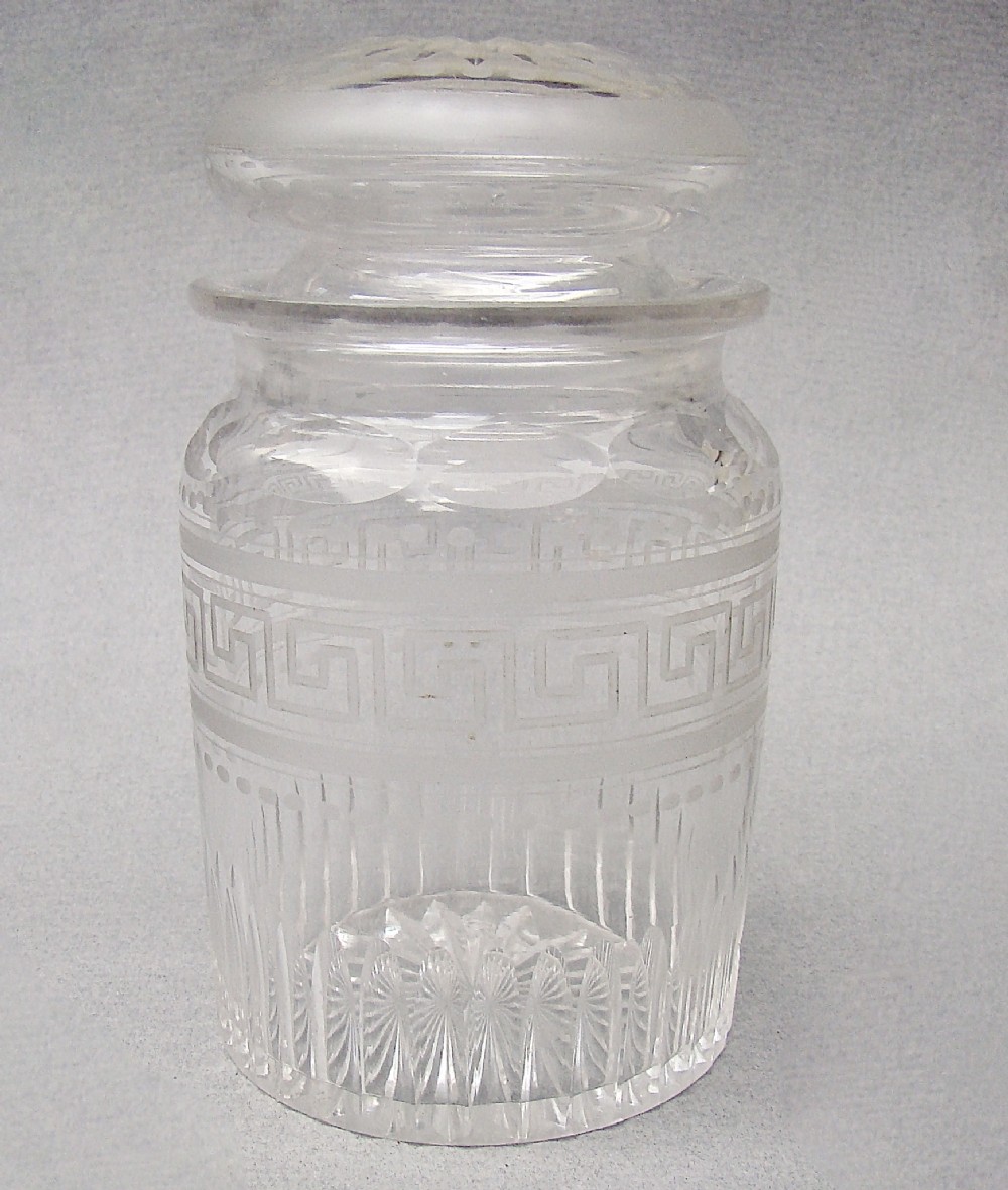 A small clear Victorian glass pickle jar with a fitted lid, decorated with an etched Greek key motif and cut glass base.