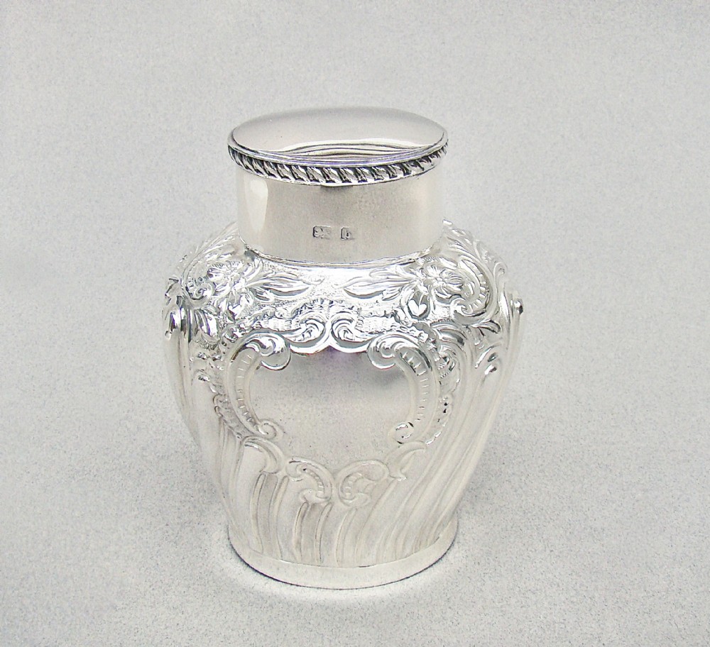 An Edwardian sterling silver tea caddy featuring repousse floral scroll decoration, a gadrooned rim lid, and British hallmarks stamped on the neck.