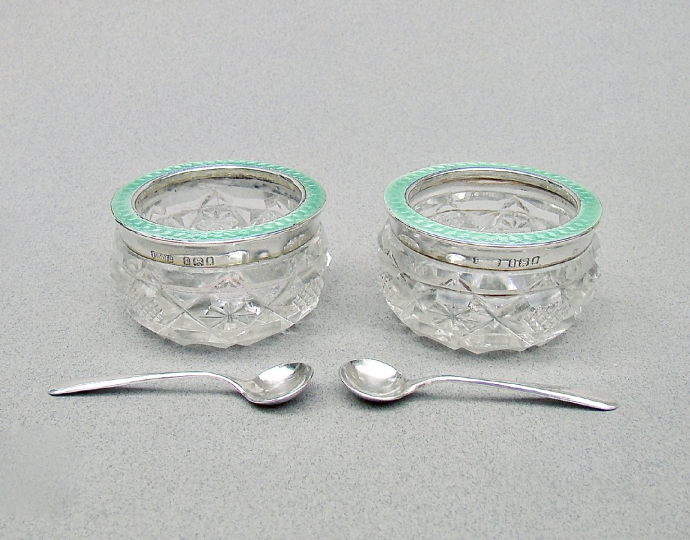 A pair of circular cut glass salt cellars featuring sterling silver rims decorated with mint green guilloche enamel, shown alongside two small silver salt spoons.