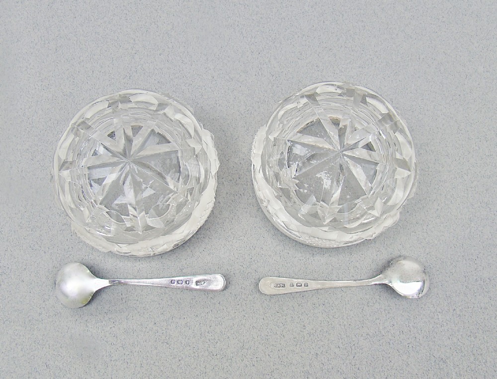 A pair of circular cut glass salt cellars featuring sterling silver rims decorated with mint green guilloche enamel, shown alongside two small silver salt spoons. View V