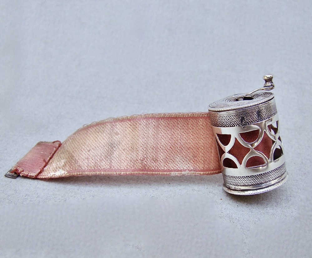 A drum-shaped 19th-century Dutch silver tape measure featuring a pierced geometric casing, top winding handle, and extending pink silk ribbon. View II