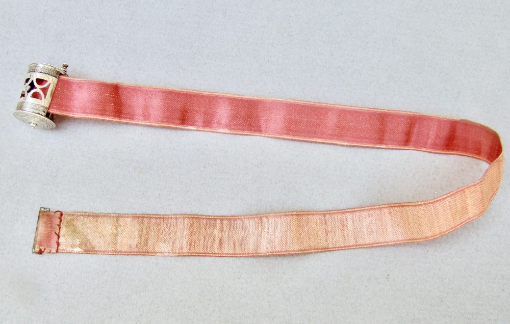 A drum-shaped 19th-century Dutch silver tape measure featuring a pierced geometric casing, top winding handle, and extending pink silk ribbon. View IV