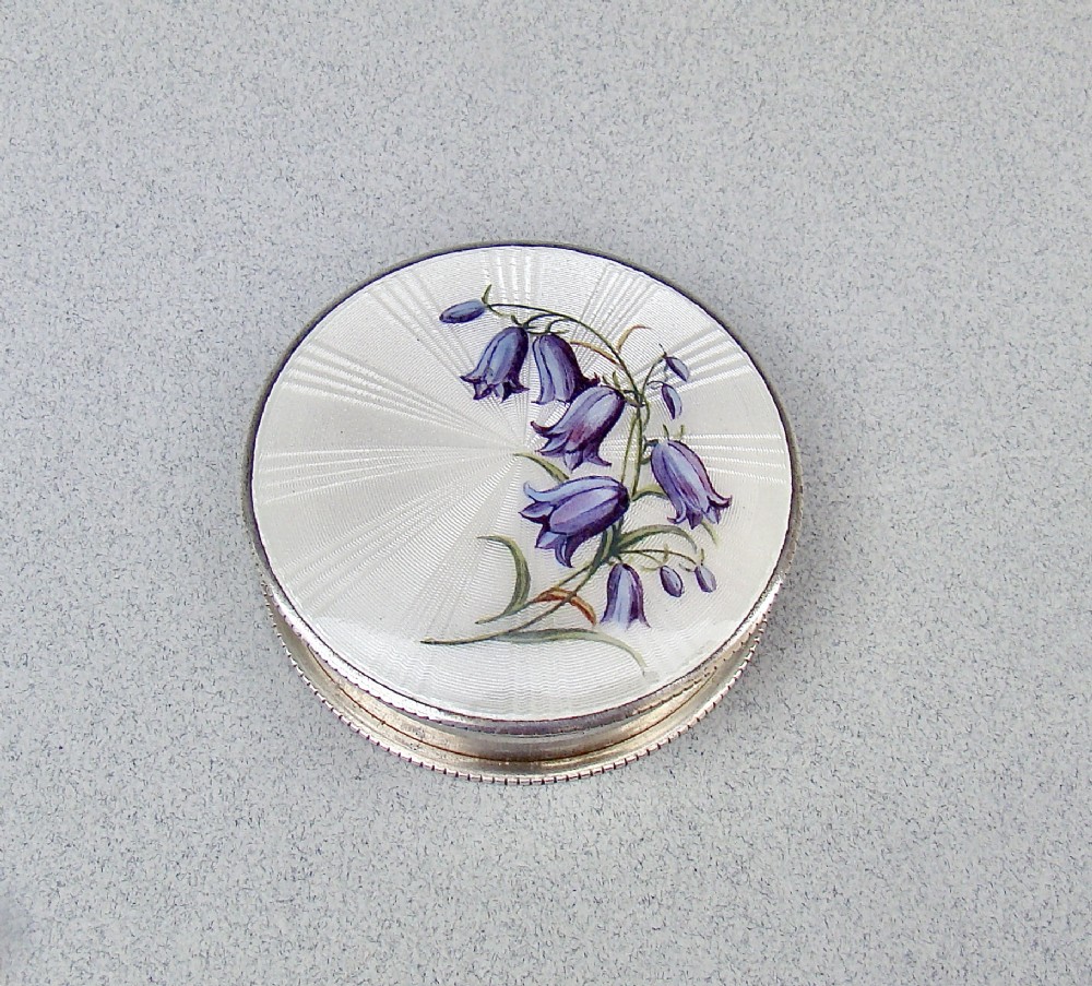 A circular Edwardian sterling silver trinket box with a white sunburst guilloche enamel lid decorated with hand-painted purple harebell flowers.