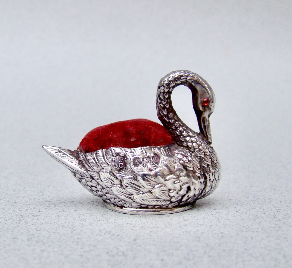 An Edwardian sterling silver swan pin cushion featuring chased feather details, a red glass eye, and a red velvet cushion insert.