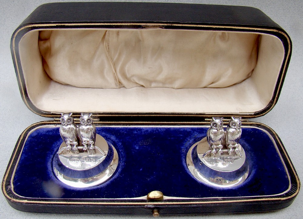 A pair of Edwardian sterling silver owl-form menu holders positioned inside a fitted black presentation case lined with blue velvet and beige silk.