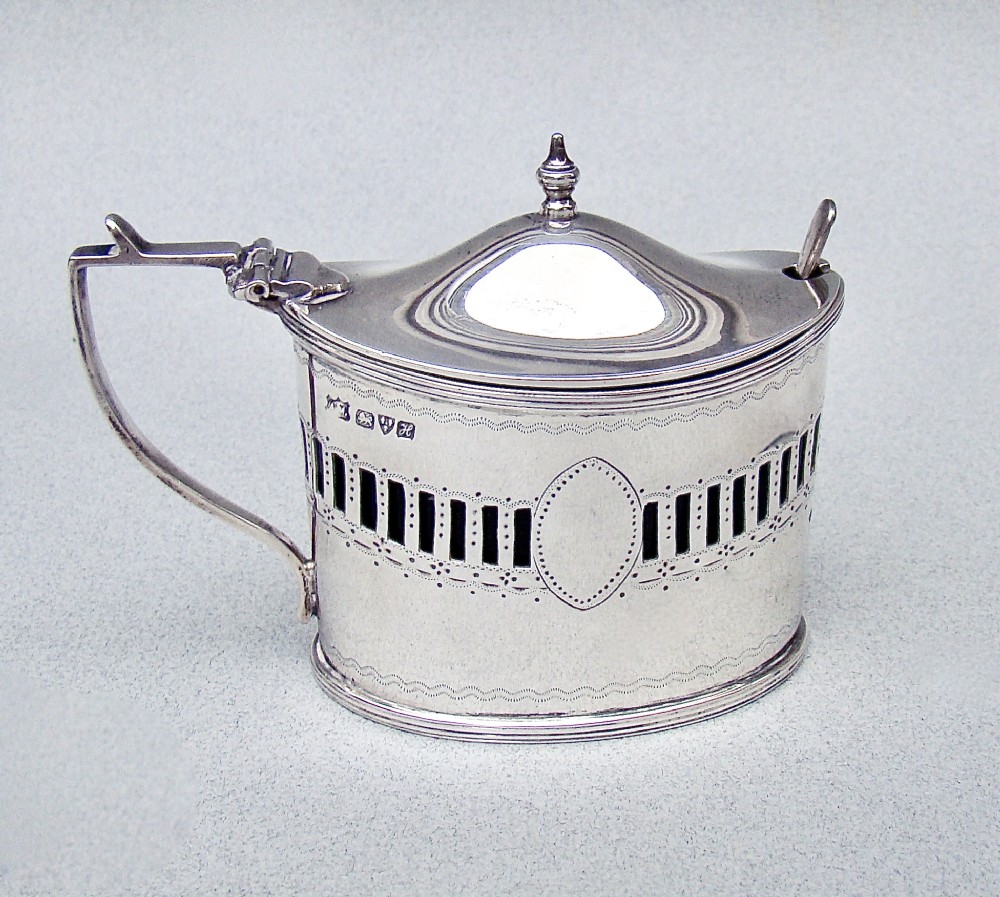 An oval Edwardian sterling silver mustard pot featuring pierced geometric fretwork, an engraved cartouche, a hinged dome lid with a finial, and a side handle.