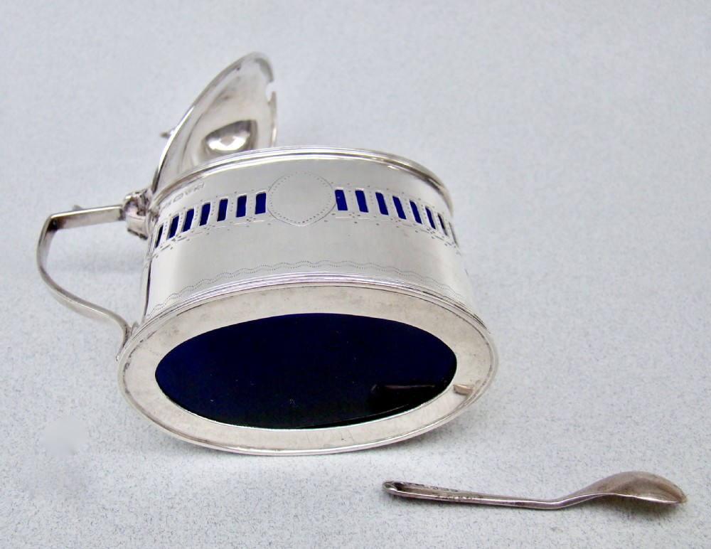 An oval Edwardian sterling silver mustard pot featuring pierced geometric fretwork, an engraved cartouche, a hinged dome lid with a finial, and a side handle. View V