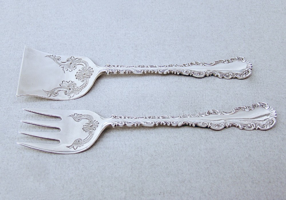 A cased set of two late Victorian sterling silver sardine servers featuring engraved scroll decorations resting inside an open fitted presentation box. View II