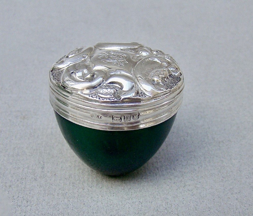 An Edwardian sterling silver pill box with a repousse lid resting on a rounded dark green glass base.